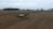 A large agricultural field with a tractor and planter in the center, preparing the ground. The scene is expansive with flat, recently tilled soil, bordered on the horizon by trees and a few farm buildings. The sky is overcast, lending a muted mood to the scene.