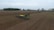 A large agricultural field with a tractor and planter in the center, preparing the ground. The scene is expansive with flat, recently tilled soil, bordered on the horizon by trees and a few farm buildings. The sky is overcast, lending a muted mood to the scene.