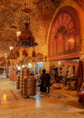 A richly decorated interior with ornate metal lanterns and arches featuring intricate patterns. The space is filled with various textile and metal objects, showcasing a traditional market setting. Several people are gathered in a group, engaging in conversation or browsing the items on display.