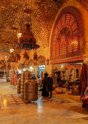 A richly decorated interior with ornate metal lanterns and arches featuring intricate patterns. The space is filled with various textile and metal objects, showcasing a traditional market setting. Several people are gathered in a group, engaging in conversation or browsing the items on display.