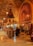 A richly decorated interior with ornate metal lanterns and arches featuring intricate patterns. The space is filled with various textile and metal objects, showcasing a traditional market setting. Several people are gathered in a group, engaging in conversation or browsing the items on display.