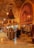 A richly decorated interior with ornate metal lanterns and arches featuring intricate patterns. The space is filled with various textile and metal objects, showcasing a traditional market setting. Several people are gathered in a group, engaging in conversation or browsing the items on display.