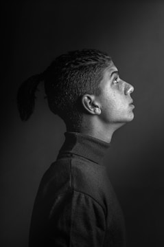 A monochrome portrait of a person in profile, featuring a sharp side view against a dark background. The individual has a short haircut with a ponytail and is wearing a turtleneck sweater. The light highlights the facial features and texture of the clothing, creating a strong contrast.