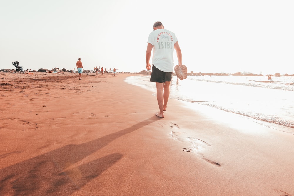 Mann in weißem T-Shirt und blauen Shorts geht tagsüber auf braunem Sand spazieren