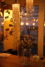 Softly lit interior with crystal chandeliers hanging above a wooden table. The table is adorned with tall glass vases containing dried flowers. The warm glow from the lights creates a cozy and inviting atmosphere. In the background, blurred outlines of chairs and other decorations are visible through large windows.