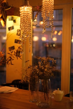 Softly lit interior with crystal chandeliers hanging above a wooden table. The table is adorned with tall glass vases containing dried flowers. The warm glow from the lights creates a cozy and inviting atmosphere. In the background, blurred outlines of chairs and other decorations are visible through large windows.