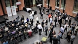 A large group of people gathered in a spacious room with brick walls, some seated in rows while others stand and engage in conversation. The atmosphere appears formal, with people dressed in business attire. There are banners with logos and numbers displayed on stands, suggesting a corporate event or conference.