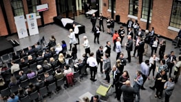 A large group of people gathered in a spacious room with brick walls, some seated in rows while others stand and engage in conversation. The atmosphere appears formal, with people dressed in business attire. There are banners with logos and numbers displayed on stands, suggesting a corporate event or conference.