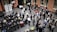 A large group of people gathered in a spacious room with brick walls, some seated in rows while others stand and engage in conversation. The atmosphere appears formal, with people dressed in business attire. There are banners with logos and numbers displayed on stands, suggesting a corporate event or conference.