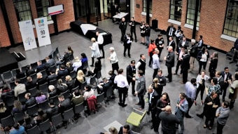 A large group of people gathered in a spacious room with brick walls, some seated in rows while others stand and engage in conversation. The atmosphere appears formal, with people dressed in business attire. There are banners with logos and numbers displayed on stands, suggesting a corporate event or conference.