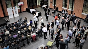 A large group of people gathered in a spacious room with brick walls, some seated in rows while others stand and engage in conversation. The atmosphere appears formal, with people dressed in business attire. There are banners with logos and numbers displayed on stands, suggesting a corporate event or conference.