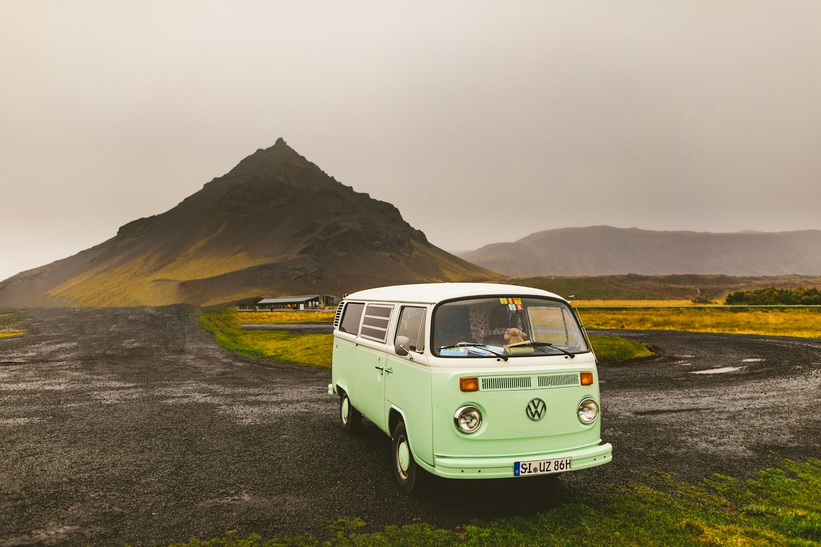 Mint-green vintage Volkswagen camper van parked on a gravel pull-off with a steep pyramid-shaped mountain and misty hills behind it in Iceland