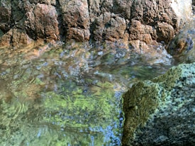 A close-up of a stream or small body of water flowing over rocks. The water appears clear, reflecting the light and revealing various shades of green, brown, and blue beneath the surface. Rough, textured rocks form a natural barrier on one side.