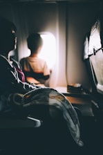 A dimly lit airplane cabin interior shows two passengers seated. A child looks out through the bright window while an adult, whose face is mostly shadowed, sits next to them. A blanket drapes across the adult's lap, and the seat's headrests are visible.