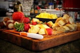 A wooden cutting board is topped with a vibrant assortment of fruits and bread. Strawberries, apple slices, and chunks of pineapple are visibly fresh and colorful. A knife rests on the board alongside sliced pieces of bread. The background showcases various kitchen items slightly out of focus, suggesting a busy and lively kitchen setting.