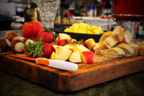 A wooden cutting board is topped with a vibrant assortment of fruits and bread. Strawberries, apple slices, and chunks of pineapple are visibly fresh and colorful. A knife rests on the board alongside sliced pieces of bread. The background showcases various kitchen items slightly out of focus, suggesting a busy and lively kitchen setting.