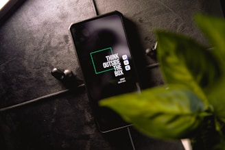 A smartphone with the phrase 'Think Outside The Box' displayed on the screen is placed on a dark textured surface. Next to the phone, a pair of black wireless earbuds can be seen. In the foreground, there is a green leafy plant.