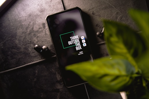 A smartphone with the phrase 'Think Outside The Box' displayed on the screen is placed on a dark textured surface. Next to the phone, a pair of black wireless earbuds can be seen. In the foreground, there is a green leafy plant.