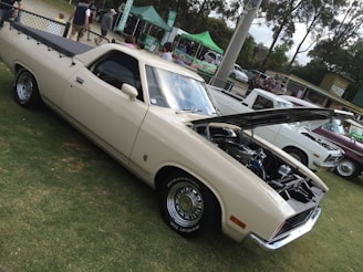 A vintage cream-colored pickup truck with its hood open is parked on grass. In the background, a community event is taking place with people gathering around stalls under green tents. Other classic cars are lined up nearby, and trees surround the area.