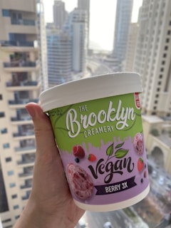 A hand is holding a tub of vegan ice cream against an urban backdrop with tall buildings. The ice cream tub is labeled 'The Brooklyn Creamery' and 'Berry 3X', with vibrant imagery of berries and a green and purple color scheme.