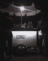 A street coffee cart illuminated by a bright light from above. The cart has a clean, modern design with the text 'Street Coffee' prominently displayed. Several coffee-making items, including syrups and bottles, are visible behind the cart. Two people are present, with one on each side of the cart, but their features are obscured by shadows.