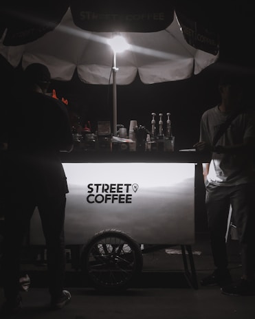 A street coffee cart illuminated by a bright light from above. The cart has a clean, modern design with the text 'Street Coffee' prominently displayed. Several coffee-making items, including syrups and bottles, are visible behind the cart. Two people are present, with one on each side of the cart, but their features are obscured by shadows.