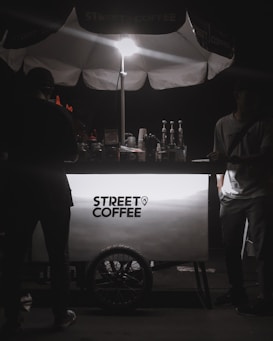 A street coffee cart illuminated by a bright light from above. The cart has a clean, modern design with the text 'Street Coffee' prominently displayed. Several coffee-making items, including syrups and bottles, are visible behind the cart. Two people are present, with one on each side of the cart, but their features are obscured by shadows.