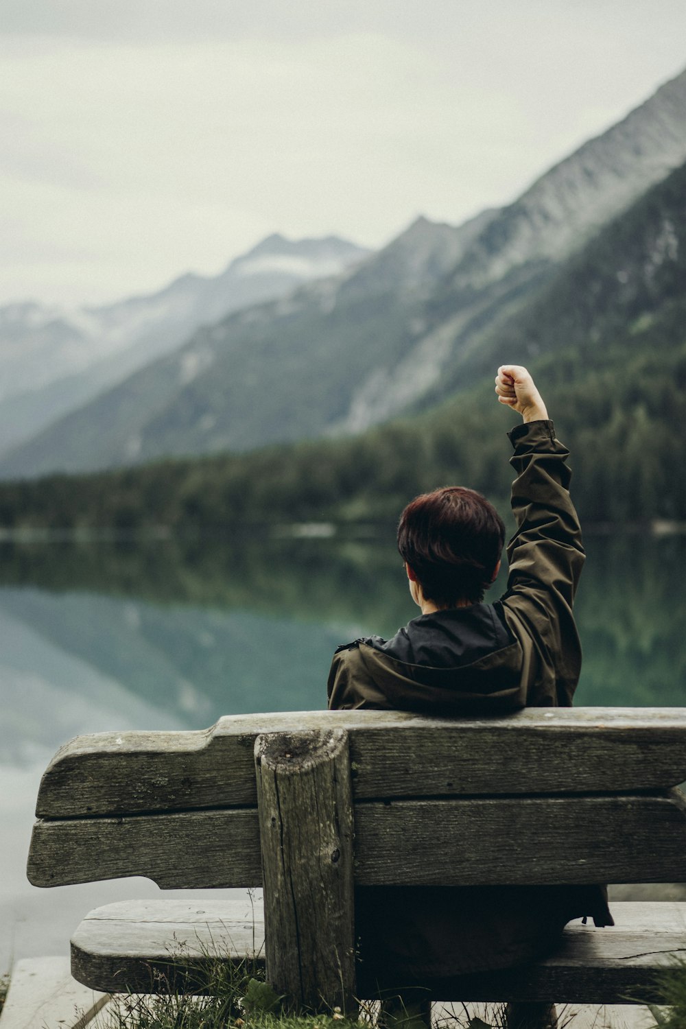 man in black and brown jacket sitting on brown wooden bench looking at lake during daytime