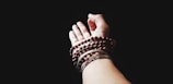 A hand is extended against a dark background, adorned with multiple strands of beaded bracelets, some in dark wood tones. The focus is on the hand and jewelry, with bright lighting contrasting against the shadowed background.