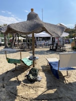 Two lounge chairs with towels are placed under a large straw parasol on a sandy beach. A backpack is on the sand in front of the chairs. In the background, there are picnic tables and a bar with people sitting around.