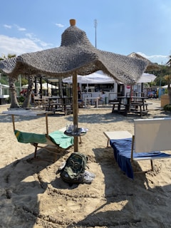 Two lounge chairs with towels are placed under a large straw parasol on a sandy beach. A backpack is on the sand in front of the chairs. In the background, there are picnic tables and a bar with people sitting around.