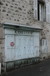 The image depicts an old building facade with a weathered blue door and the word 'Coiffeur' written above it, indicating a hairdresser. The walls are made of rough, gray stone, and there is a closed window with wooden shutters beside the door. A red sign near the door warns against parking.
