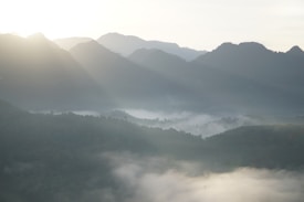 A serene mountain landscape is enveloped in mist, with sunlight streaming through gaps in the mountain range, creating a soft, diffused light. The mountains form a series of peaks and valleys, gradually fading into the distance. The scene exudes tranquility and natural beauty.