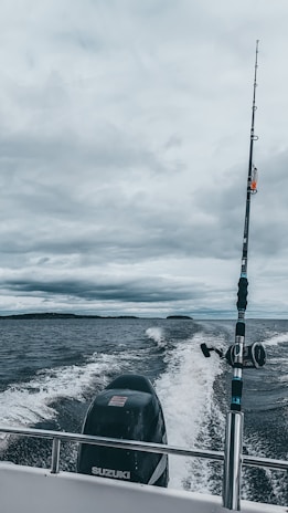 A fishing rod stands upright on a moving boat against a backdrop of cloudy skies and expansive water. The boat motor leaves a trail of white wake behind as it traverses the dark, choppy waters.