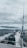 A fishing rod stands upright on a moving boat against a backdrop of cloudy skies and expansive water. The boat motor leaves a trail of white wake behind as it traverses the dark, choppy waters.