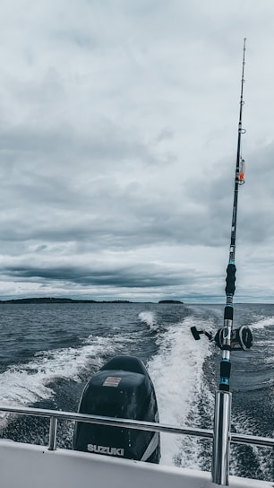 A fishing rod stands upright on a moving boat against a backdrop of cloudy skies and expansive water. The boat motor leaves a trail of white wake behind as it traverses the dark, choppy waters.