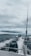 A fishing rod stands upright on a moving boat against a backdrop of cloudy skies and expansive water. The boat motor leaves a trail of white wake behind as it traverses the dark, choppy waters.