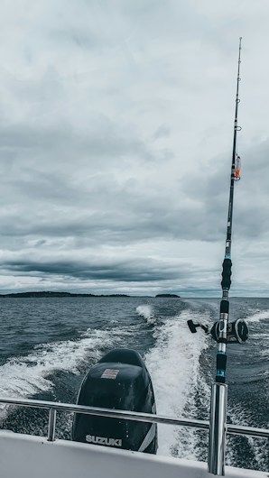 A fishing rod stands upright on a moving boat against a backdrop of cloudy skies and expansive water. The boat motor leaves a trail of white wake behind as it traverses the dark, choppy waters.