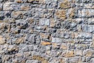 A close-up view of a textured stone wall, with irregularly shaped stones in varying shades of gray and brown, meticulously fitted together to form a solid structure.