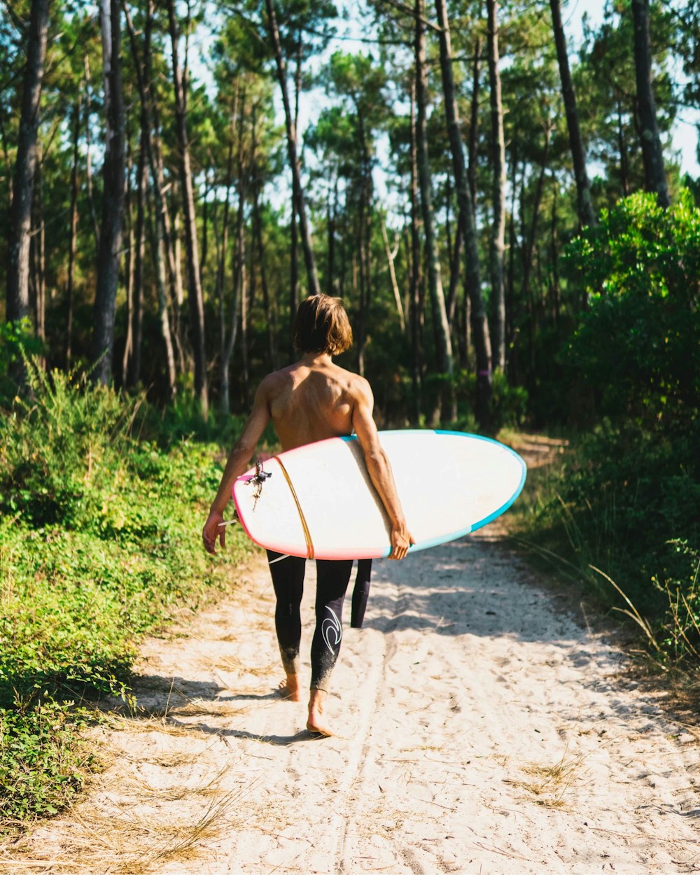 mulher no biquíni preto e branco segurando a prancha de surf branca andando na estrada de terra durante o dia