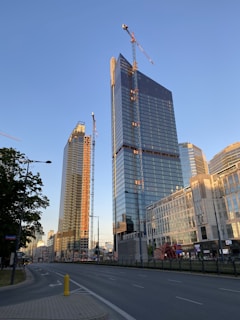 Several tall buildings are under construction with cranes visible at their peaks. The structures have a modern architectural design with large glass windows reflecting sunlight. The street in the foreground is empty with trees lining one side.