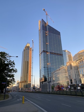 Several tall buildings are under construction with cranes visible at their peaks. The structures have a modern architectural design with large glass windows reflecting sunlight. The street in the foreground is empty with trees lining one side.