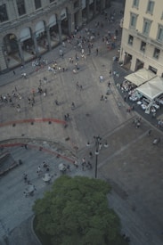An aerial view of a busy plaza surrounded by historic buildings. The scene includes numerous people walking across the stone pavement, some gathered near storefronts or outdoor dining areas with white umbrellas. A section of green trees is visible in the lower portion, and multiple outdoor seating areas with round tables are scattered throughout the plaza.