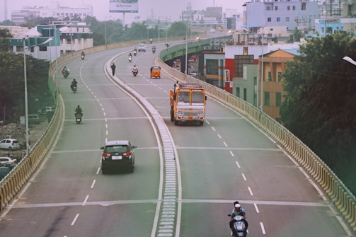 A curved urban roadway with light traffic includes several vehicles such as cars, motorcycles, a bus, and an auto-rickshaw. Buildings line the background, indicating a city setting, while the surrounding greenery contrasts with the concrete structures.