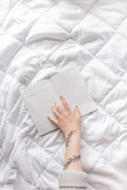 A hand with a tattoo of leaves rests on an open notebook with handwritten notes, placed on a soft, white quilted surface.