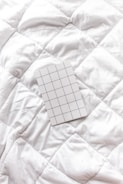 A white grid-patterned notepad rests on a textured white quilted surface. The pattern on the notepad consists of evenly spaced blue lines forming squares.