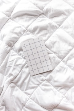 A white grid-patterned notepad rests on a textured white quilted surface. The pattern on the notepad consists of evenly spaced blue lines forming squares.