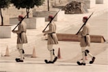 Three individuals dressed in traditional military uniforms march in a synchronized manner. They carry rifles over their shoulders and wear beige kilts, white leggings, and red berets. The setting is an outdoor area with stone walls, trees, and several orange traffic cones.