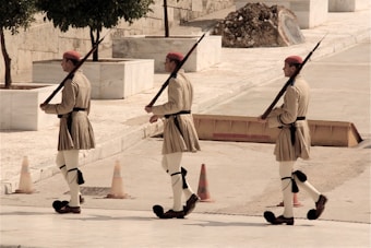 Three individuals dressed in traditional military uniforms march in a synchronized manner. They carry rifles over their shoulders and wear beige kilts, white leggings, and red berets. The setting is an outdoor area with stone walls, trees, and several orange traffic cones.