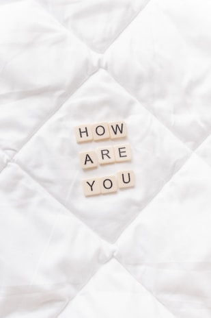 Letter tiles spell out 'HOW ARE YOU' placed neatly on a soft, white quilted surface.